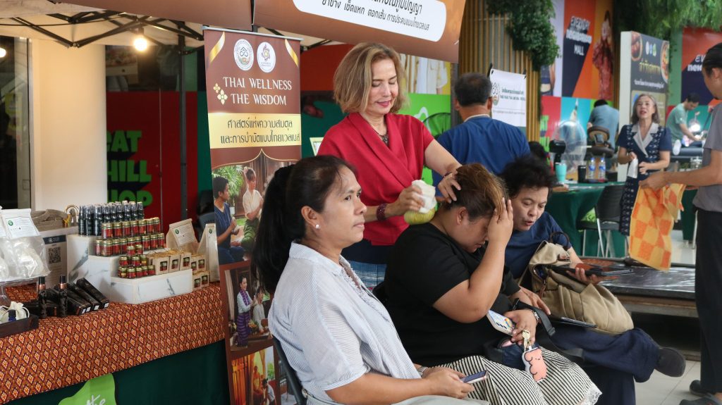 Thai wellness booth at a market-style expo; a woman in red massages a seated client while others sit nearby and browse products.