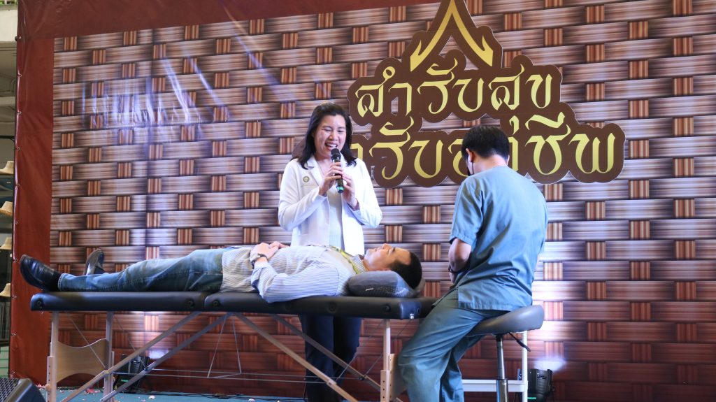 Healthcare training scene: a woman in a white coat speaks into a microphone beside a patient lying on an examination/treatment table, with a second clinician in scrubs nearby, against a patterned backdrop with gold lettering.