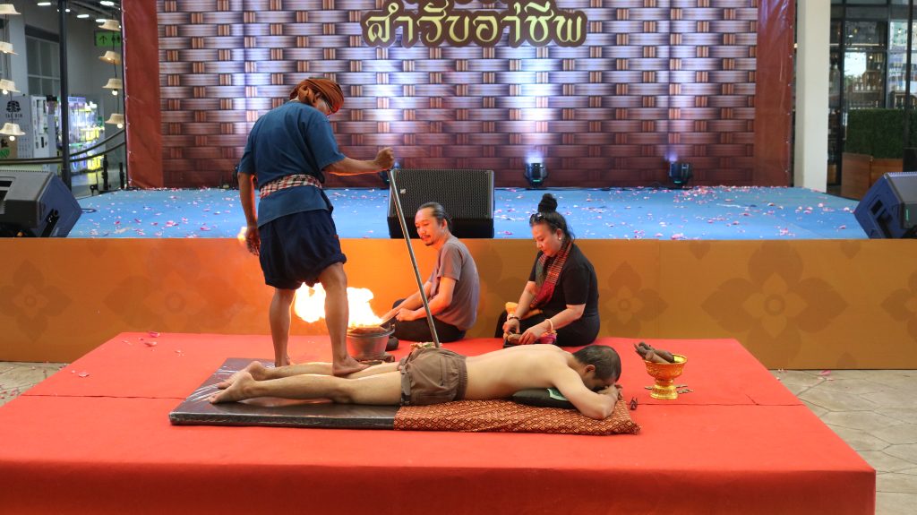 Stage scene of a fire-walking ritual: a man in a blue outfit with a headband stands over a glowing fire pit holding a staff, while two participants sit nearby on the mat and watch; one person lies prone on the mat, with a decorative backdrop and scattered petals around the area.