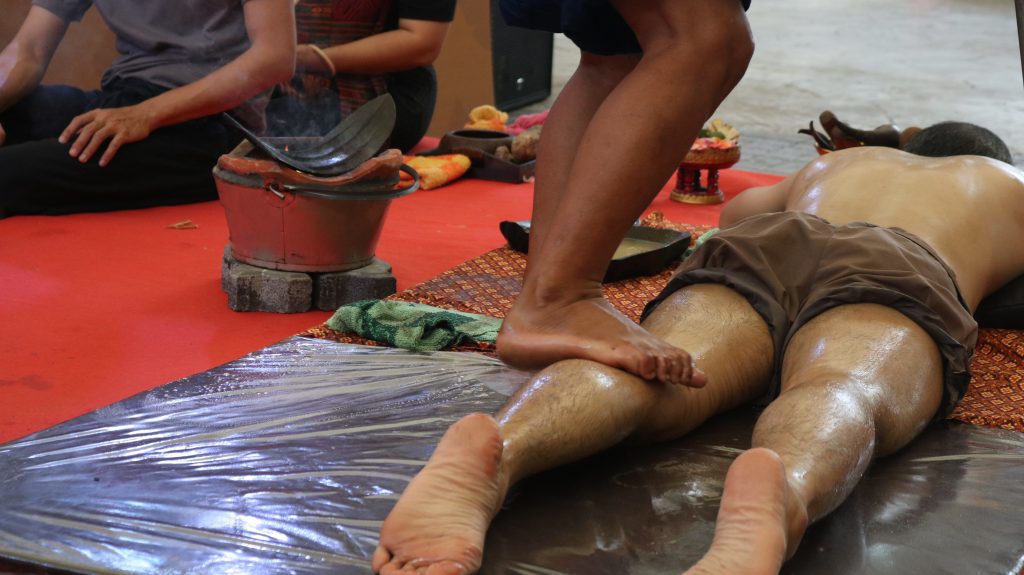 Person lying face down on a red mat during a massage, with a practitioner nearby and a heated pot on bricks in the background