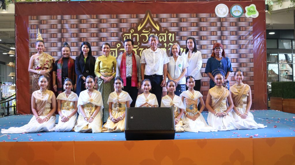 Group of performers in traditional Thai attire posing on a stage with a woven backdrop and event banners behind them.