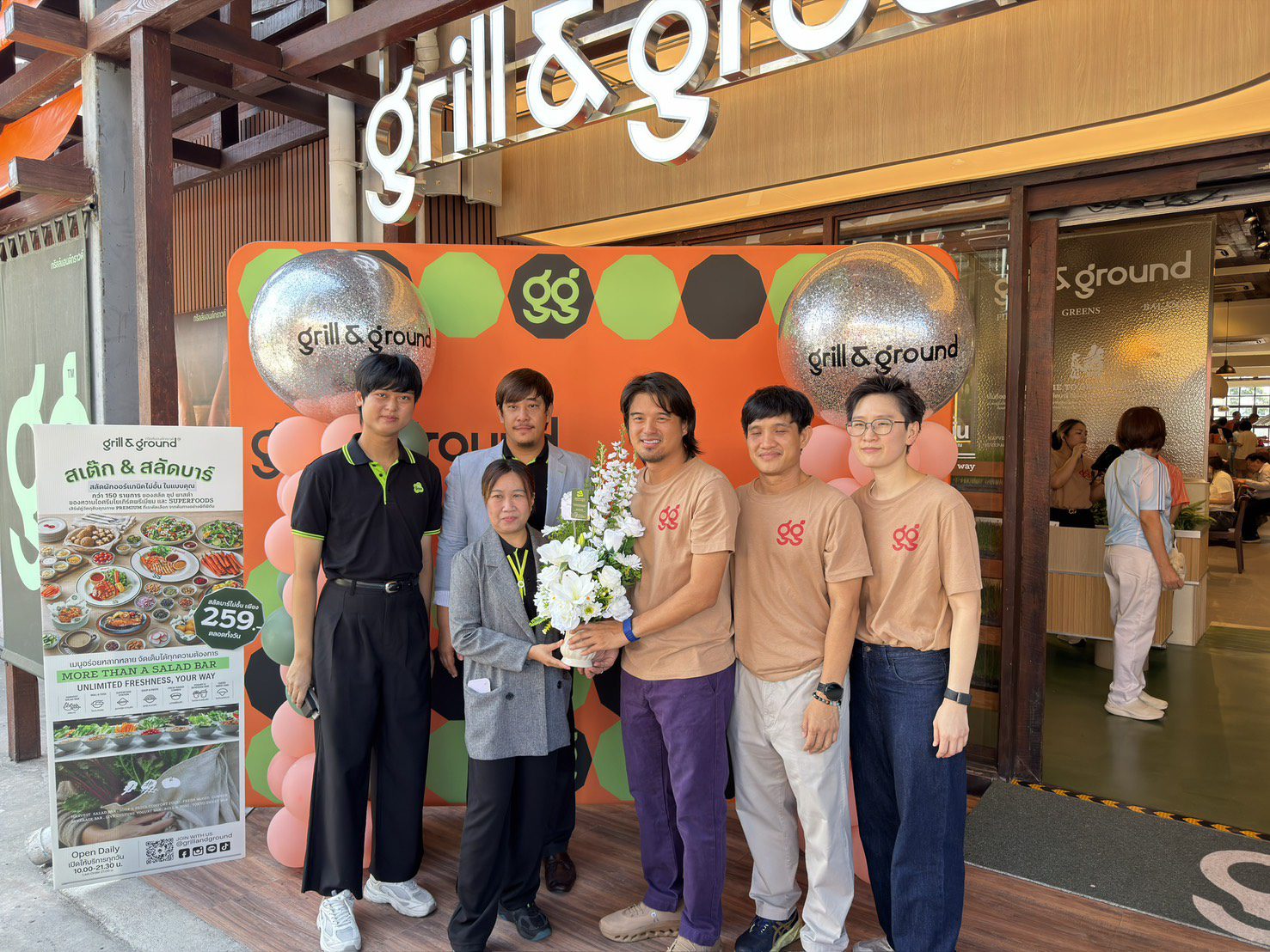 Six people posing for a photo in front of a bright orange grill & ground backdrop outside a restaurant; one person holds a white bouquet of flowers.